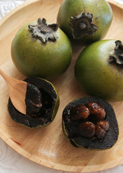 a wooden plate holds four ripe Wilson Black Sapote fruits - one fruit is cut in half exposing dark, soft flesh